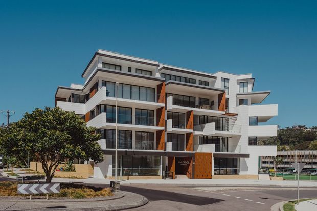 A contemporary multi-storey apartment building viewed from the street, featuring stepped and cantilevered white balconies, extensive floor-to-ceiling glazing, dark window frames, and warm orange-brown vertical facade panels. The building sits on an urban corner with landscaped trees, paved frontage, and adjacent residential development under a clear blue sky.