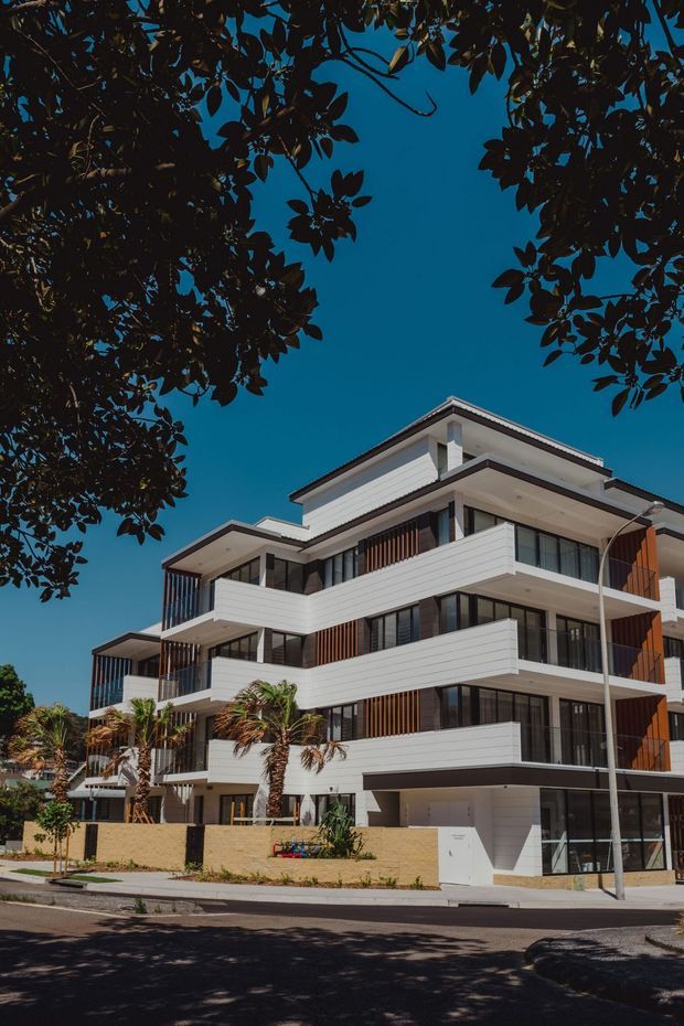 Street-level exterior photograph of a contemporary multi-storey residential apartment building. The building has layered projecting balconies, white horizontal cladding, dark-framed glazing, vertical timber-look screening, and a flat roof. Palm plantings and mature tree branches frame the facade, with a paved street and sidewalk in the foreground.