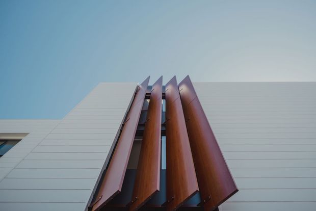Low-angle architectural exterior view of a contemporary building facade. Broad pale horizontal cladding panels frame a dramatic vertical group of projecting reddish-brown metal fins or screens, with narrow glazing visible between them. The composition emphasizes geometric repetition, clean lines, and the building's upward-reaching facade against a clear blue sky.