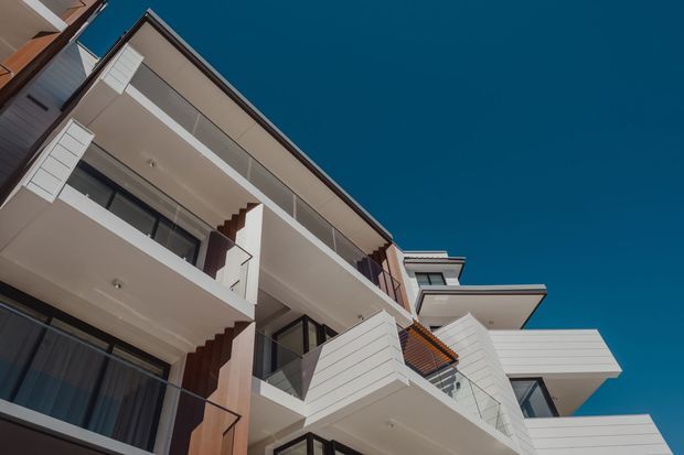 Low-angle exterior photograph of a multi-storey contemporary residential building with projecting balconies, white horizontal cladding, dark-framed glazing, glass balustrades, and angular roof and balcony forms against a clear deep-blue sky.