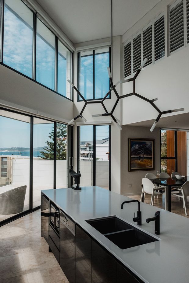 A bright, double-height contemporary kitchen and dining space with expansive black-framed glazing overlooking a coastal residential setting. A large island with a pale stone benchtop, dark integrated cabinetry, twin black sinks and sculptural black tapware dominates the foreground. The room features polished pale flooring, white walls, plantation shutters, a geometric branching pendant light, and a dining area beyond.
