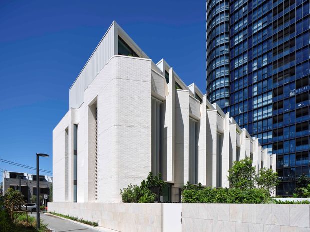 Exterior architectural photograph of a contemporary multi-storey white brick building with a rhythmic series of tall projecting fins, recessed openings, and steep angular roof forms. A landscaped forecourt and pale stone boundary wall sit in front, while a tall glazed high-rise forms the urban backdrop. Clear blue sky and strong sunlight create a crisp, high-contrast presentation.