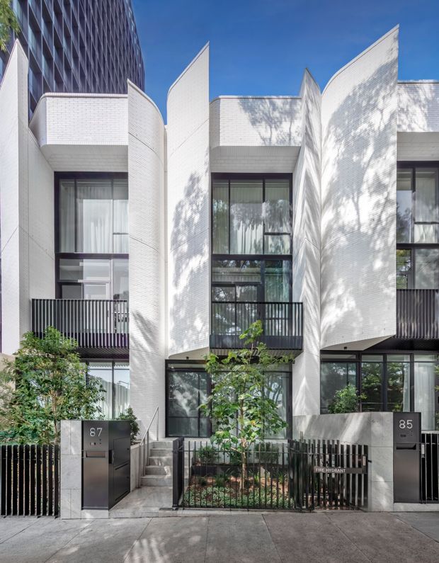 Street-level exterior view of a contemporary multi-storey residential development with three narrow townhouse-like facades. The building features white small-format brick cladding, sculptural curved and folded vertical wall elements, dark-framed floor-to-ceiling windows, black metal balcony and boundary railings, landscaped front gardens, and recessed entry doors. A taller dark-glazed building is visible behind the development.