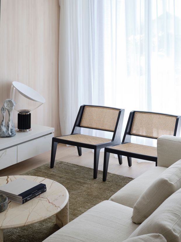 Bright, refined living room with two black-framed cane lounge chairs positioned beside a large sheer-curtained window. A cream sectional sofa, pale marble-patterned coffee table, textured neutral rug, white low media cabinet, sculptural decor and table lamp create a warm, minimalist interior.