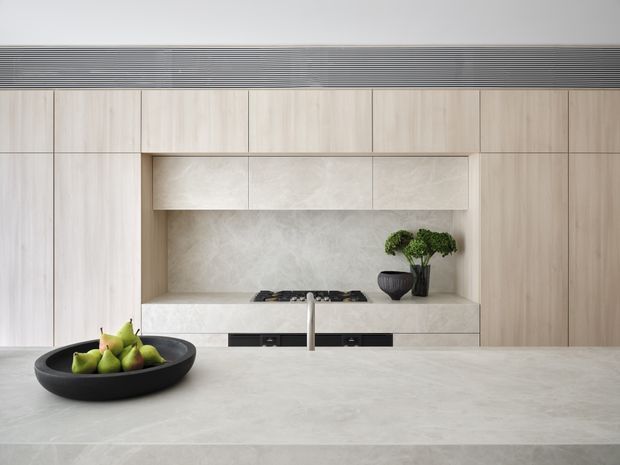 Minimalist contemporary kitchen with a large pale stone island in the foreground, light timber-look full-height cabinetry, an integrated stone backsplash and rangehood surround, and a black cooktop with a slim central tap. A black fruit bowl and small potted plants provide restrained accents. A linear dark ventilation grille runs above the cabinetry.