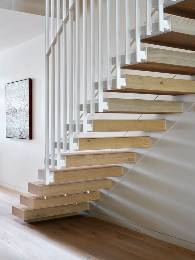 A bright, minimalist interior photograph focused on a floating staircase. Thick natural timber treads are cantilevered from a wall, paired with slender white vertical balusters and white structural supports. The staircase rises beside a smooth white wall, with dark-to-mid brown timber flooring below and a simple abstract wall artwork nearby.