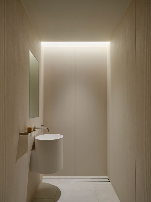 A narrow, minimalist powder room with warm beige vertically textured wall panels, pale tiled flooring, a wall-mounted cylindrical basin, slim wall-mounted tapware, and a tall frameless mirror. Concealed linear lighting creates a soft illuminated recess at the rear of the room.