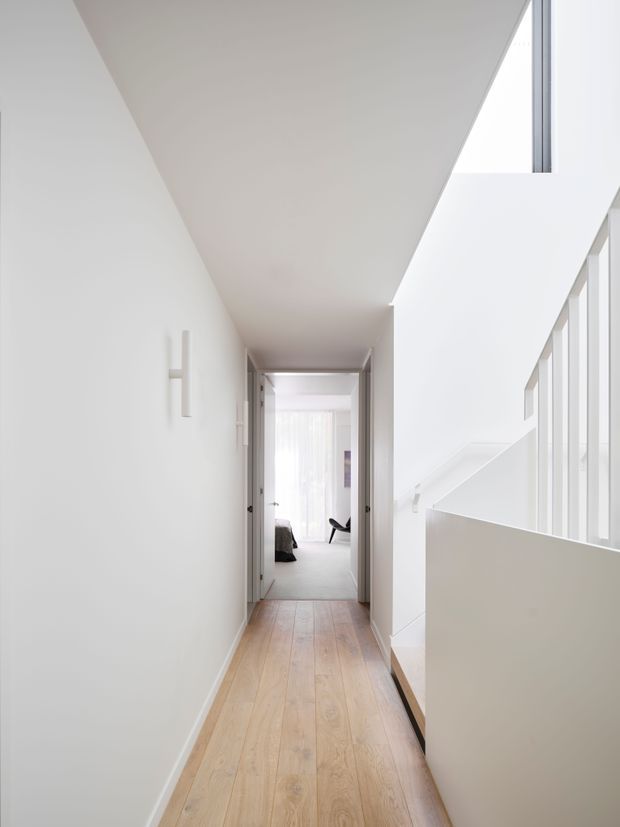 A bright, minimalist interior hallway with pale timber flooring and white walls, leading toward a bedroom. A modern open stairwell with white balustrade and tall vertical glazing occupies the right side, creating an airy, light-filled circulation space.