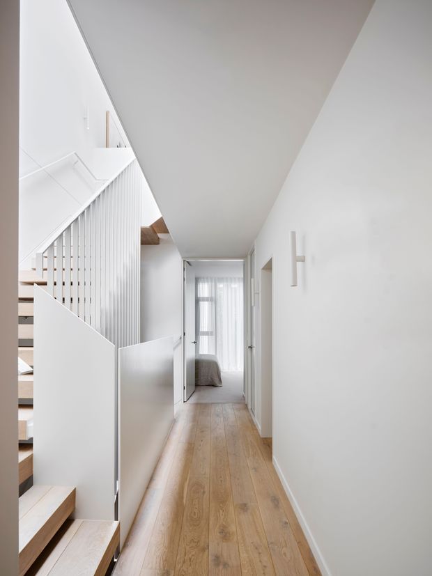A bright, narrow residential hallway with warm timber flooring, white walls, and a sculptural open staircase on the left. The staircase has white vertical balustrades and pale timber treads, with concealed storage or panelling beneath. A bedroom is visible at the end of the corridor through an open doorway, and soft daylight filters through sheer curtains. The overall design is clean, minimalist, and contemporary.