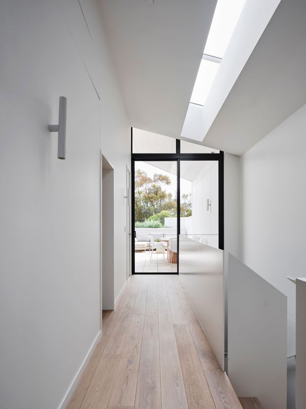 A bright, minimalist hallway or landing with pale timber flooring, white walls, a black-framed glazed door, and a long linear skylight. The corridor leads to a light-filled outdoor terrace or courtyard, with a stair opening and simple wall-mounted cylindrical light visible in the foreground.