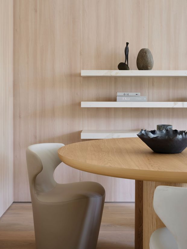 Refined dining area featuring a round light-oak dining table with sculptural upholstered chairs, set against a full-height pale timber wall. Three slim white stone shelves display understated ceramics and sculptural objects, creating a warm, minimalist composition.