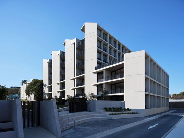 Exterior view of a contemporary multi-storey apartment building with a long rectilinear form, repeated balconies, white rendered or concrete-looking walls, dark metal balcony balustrades, and a landscaped stepped entry. The building fronts a paved driveway or access road under a clear blue sky.