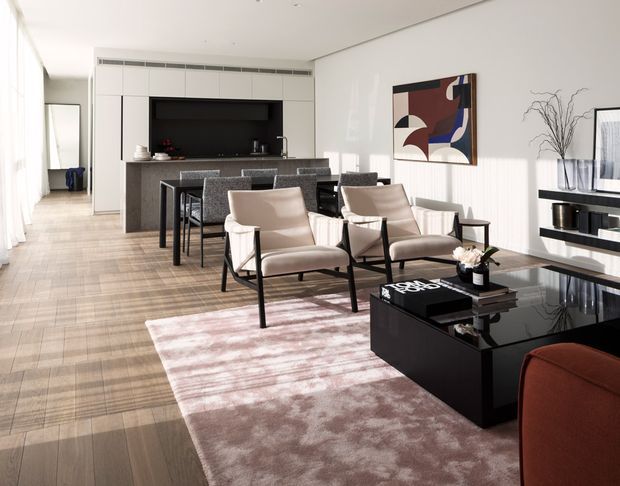 Bright open-plan contemporary living and dining area with a kitchen at the rear. The room features pale timber or stone-look flooring, white walls and ceiling, a dark stone kitchen island, black built-in shelving and coffee table, upholstered dining chairs, sculptural lounge chairs, and a muted pink patterned rug. Large sheer-curtained glazing brings in abundant natural light, while abstract artwork adds a graphic accent.