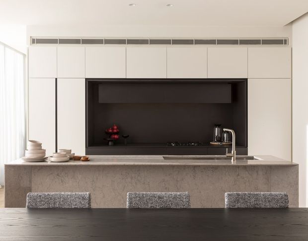 Contemporary open-plan kitchen viewed across a dark dining table, with a large stone island featuring three upholstered stools. The kitchen has full-height pale cabinetry, a dramatic dark recessed cooking and splashback zone, integrated ventilation, a minimalist cooktop, and a brushed metal tap beside the sink. Soft daylight enters from the left through sheer curtains, creating a refined, neutral interior.