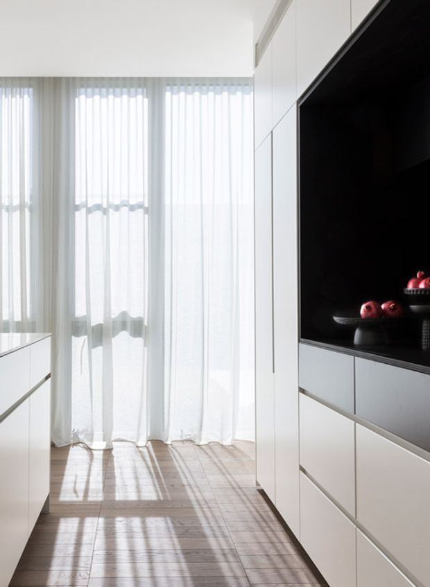 A bright, minimalist interior with full-height sheer curtains filtering sunlight onto a light timber floor. White built-in cabinetry runs along the right wall and includes a large black recessed display or media niche with decorative red objects. Additional sleek white joinery is visible on the left, creating a clean, streamlined corridor-like space.