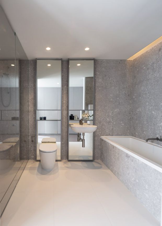 A contemporary bathroom featuring a freestanding toilet, wall-mounted basin with mirrored cabinetry, a built-in bathtub, large-format grey stone-look wall surfaces, and pale tiled flooring. Recessed ceiling lights and concealed warm LED lighting add a refined, softly illuminated atmosphere. A glass-enclosed shower is visible on the left.