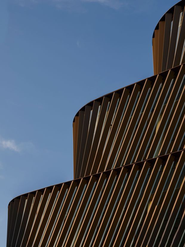 Upward-looking architectural detail of a contemporary multi-storey building facade. The dark brown and bronze-toned exterior is composed of repeated vertical fins or louvres, with stepped curved building volumes and narrow glazing between the fins, set against a clear blue sky.