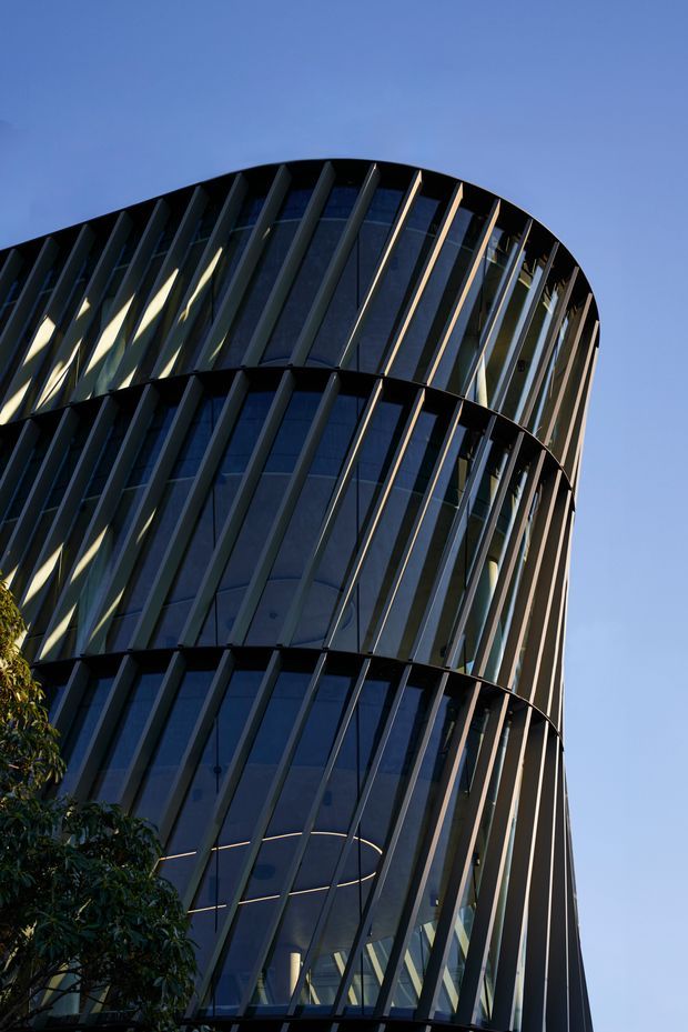 Low-angle exterior view of a tall contemporary commercial building with a rounded, curving form, dark reflective curtain-wall glazing, and repeated diagonal vertical fins. The stacked curved facade elements create a strong sculptural rhythm against a clear blue sky, with a small amount of tree foliage at the lower edge.