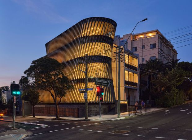 Dusk exterior view of a prominent multi-storey urban building at a street intersection. The main structure has a sculptural, curving facade wrapped in closely spaced vertical fins, with warm interior lighting visible through the glazing. An adjoining taller traditional building and street trees, traffic lights, road markings, and signage frame the scene.