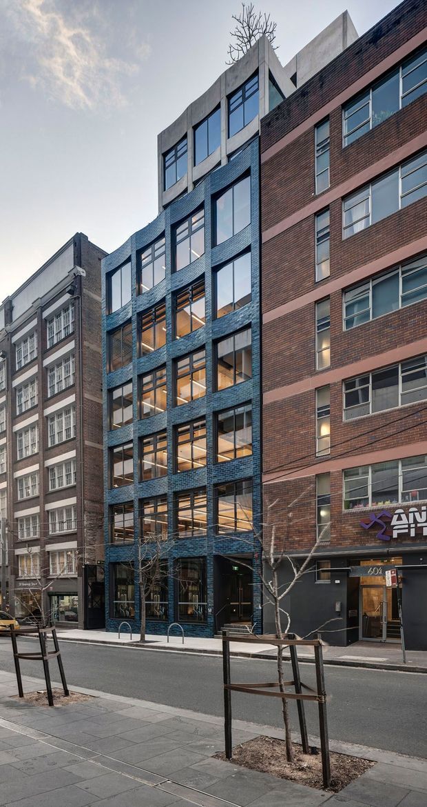 Street-level view of a narrow contemporary urban commercial building inserted between older brick buildings. The main facade uses dark blue glazed brick, large vertically proportioned windows, and curved projecting brick bays, with a sculptural concrete parapet and rooftop tree. Adjacent red-brick multi-storey buildings, shopfronts, street trees, paving, and bicycle rails establish a dense city streetscape.