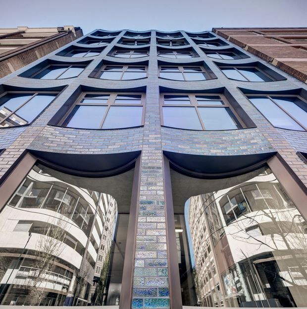 Upward-looking architectural photograph of a multi-storey urban building facade. The facade features narrow multicoloured brickwork, deep dark-framed window bays, curved projecting brick forms, and large reflective glazed openings at street level. Adjacent high-rise buildings are reflected in the glazing and visible at the edges, emphasizing the dense city setting.