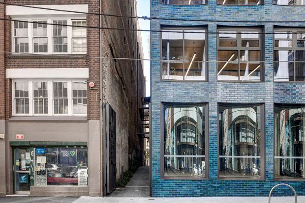 Street-level exterior view of a narrow urban laneway framed by two adjoining multi-storey buildings. The right-hand building has a distinctive blue and grey brick facade with large black-framed windows and reflective glazing, while the older building on the left features red brick, rendered panels, traditional multi-pane windows, and a ground-floor shopfront. Overhead utility cables cross the street frontage.