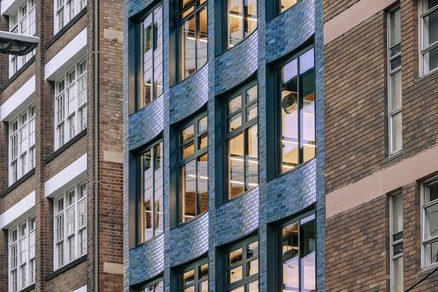 Close-up exterior view of a multi-storey urban building facade, featuring a curved central bay clad in blue glazed brick or tile, dark metal-framed windows, and adjoining brown brick elevations with white horizontal detailing. Warm interior lighting is visible through the glazing.
