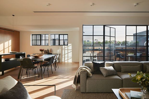 A bright, open-plan contemporary interior combining a living room, dining area and compact kitchen. Large black-framed steel-look glazing opens to a balcony or terrace, while warm timber flooring, pale walls and abundant sunlight create a spacious atmosphere. The room contains a large neutral sectional sofa, round dining table with dark chairs, and streamlined dark kitchen cabinetry. Exposed brickwork and greenery are visible beyond the glazing.
