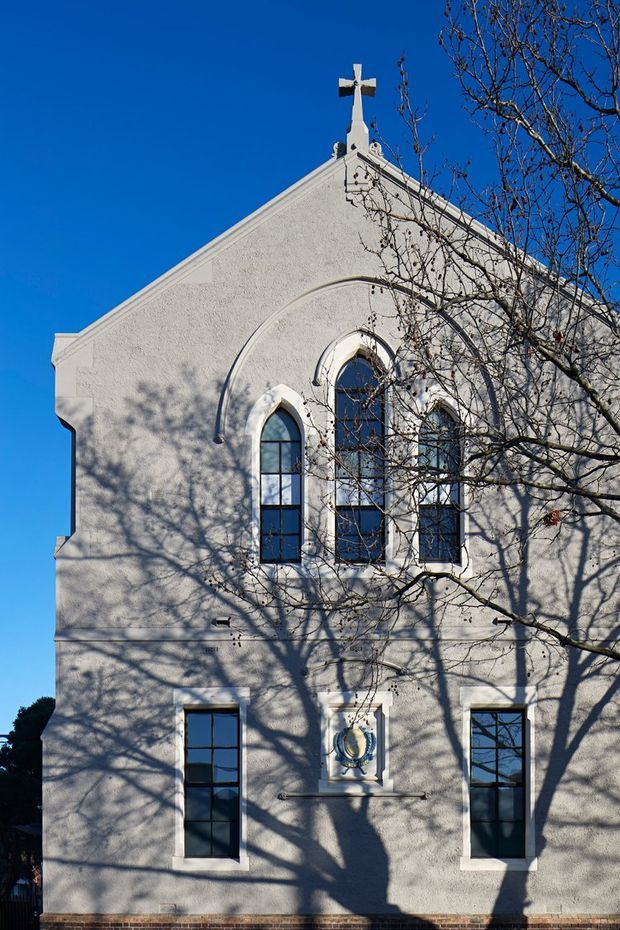Exterior view of a historic-looking white stucco church facade under a clear blue sky. The symmetrical elevation features a steep triangular gable topped with a cross, tall narrow arched windows with dark frames, decorative arched moulding, and a small ornamental relief above the lower entrance area. Leafless tree branches cast strong shadows across the facade, with a brick base visible along the bottom.
