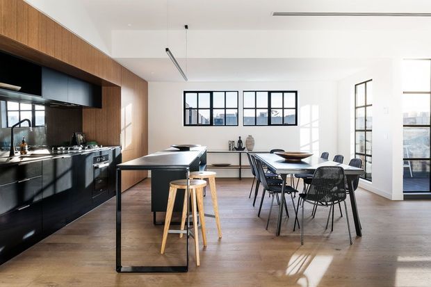 A bright, open-plan contemporary kitchen and dining area. The kitchen features matte black cabinetry, a dark stone-like benchtop, integrated appliances, timber wall panelling, and a slim island with timber stools. A black-framed dining table and chairs sit beside large industrial-style gridded windows and glazed doors, with warm timber flooring throughout. Strong natural light creates a refined, high-contrast interior.