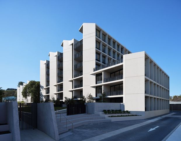 Exterior view of a contemporary multi-storey apartment building with a pale rendered or panelled facade, repeated balconies, dark metal balustrades, vertical fins, and a prominent projecting upper-level volume. The building fronts a paved street with landscaped entry steps and low boundary walls under a clear blue sky.