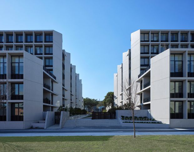 Exterior view of a contemporary multi-storey apartment development arranged as two parallel building wings around a landscaped pedestrian forecourt. The buildings feature crisp white rendered or panelled facades, extensive dark-framed glazing, recessed balconies, projecting volumes, and a formal paved entry with steps and low planting. The image is photographed from across a lawn under clear blue sky.