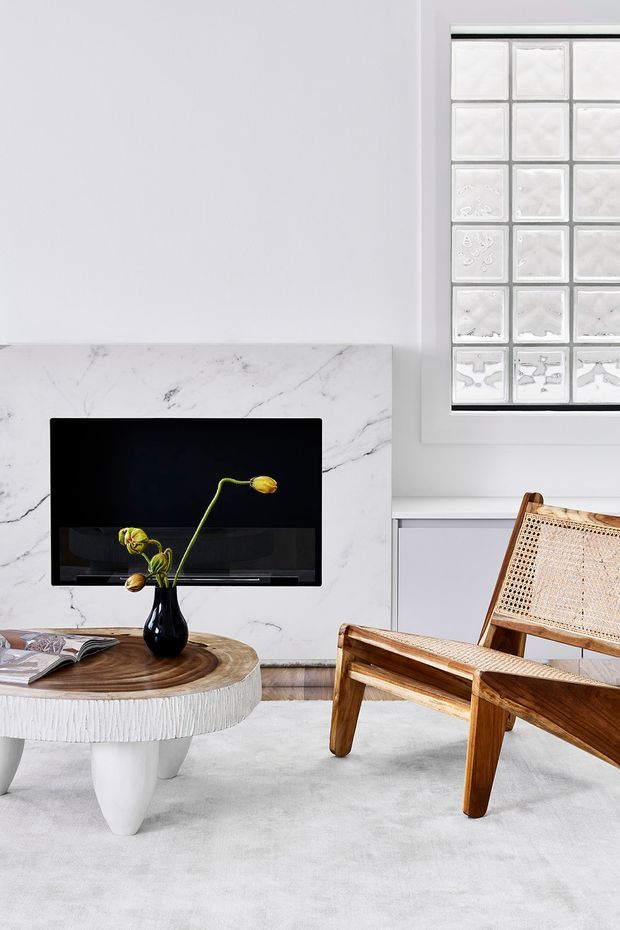 Bright, refined living room with a wall-mounted television set against a white veined marble feature wall. A sculptural round coffee table with a timber top and textured white base sits on a pale rug, accompanied by warm timber and woven cane lounge chairs. A textured glass-block window adds architectural interest.