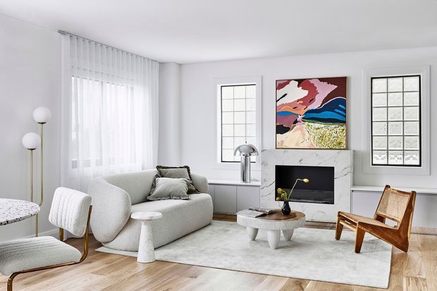 Bright, refined living room with a softly curved light-grey sofa, sculptural white coffee table, pale rug, arched lounge chair, slim globe floor lamps, and a compact television set into a marble-look fireplace surround. White walls, tall divided-light windows, sheer curtains, light timber flooring, and colourful abstract artwork create an airy contemporary setting.