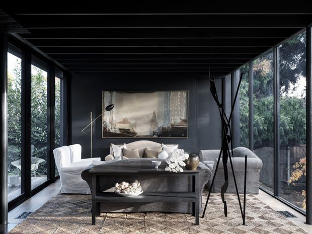 A moody, refined living room enclosed by extensive floor-to-ceiling glazing on three sides. Dark walls, ceiling beams, and window frames contrast with pale upholstered sofas, a black rectangular coffee table, a large patterned rug, sculptural decor, and abstract wall art. Dense greenery is visible outside through the glazing.