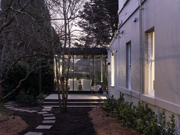 Exterior view of a contemporary home beside a landscaped garden, showing a pale rendered wall with tall windows and a glazed sunroom or enclosed terrace projecting into the garden. A timber deck with steps, stepping-stone path, dense planting, and minimal outdoor seating create a quiet, integrated indoor–outdoor setting in subdued evening light.