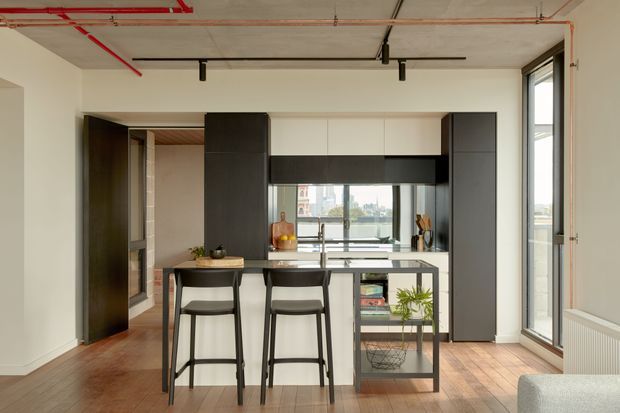 A contemporary open-plan kitchen with a compact island and two bar stools, dark and white built-in cabinetry, a black-framed window, and exposed concrete ceiling services with track lighting. Warm timber flooring contrasts with the monochrome joinery. A glazed opening at the rear provides city and waterfront views, while a living area is partially visible at the right edge.
