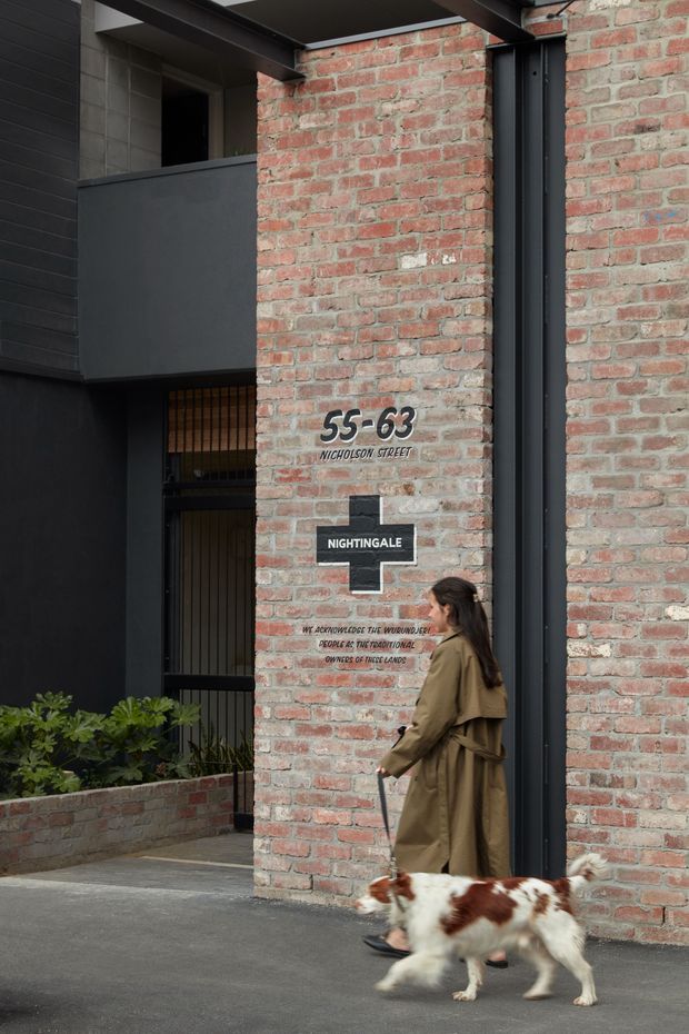 Street-level view of a contemporary building entrance or facade, featuring exposed red brick walls, dark metal cladding and framing, a recessed gated entry, and a prominent address and building sign. A pedestrian walking a dog is present in the foreground.