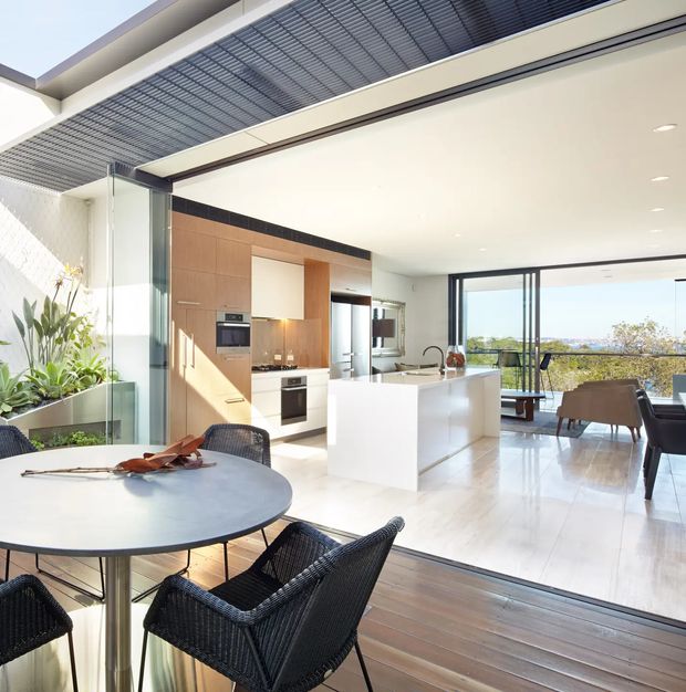 Bright open-plan kitchen and dining area with a white island, warm timber cabinetry, integrated appliances, glossy light flooring, and expansive floor-to-ceiling glazing opening toward a balcony or terrace. A round dining table with dark woven chairs occupies the foreground, while a louvred overhead structure and planted glazed lightwell add architectural interest.