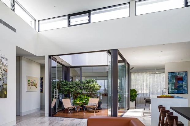 Bright contemporary open-plan interior featuring a double-height void, extensive clerestory glazing, and a glazed internal courtyard or sunroom filled with plants. The space includes a polished light-toned floor, white walls, a kitchen or bar area with stools, lounge seating, and abundant natural light.