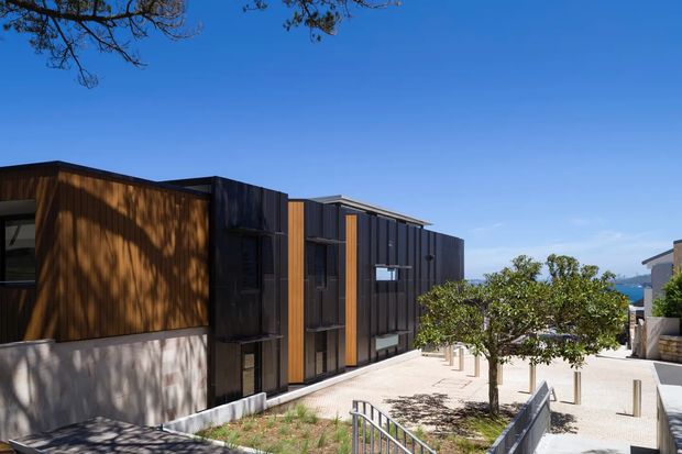 Exterior view of a contemporary multi-level building with dark vertical cladding, warm timber wall panels, extensive glazing, and a flat roofline. The building fronts a paved pedestrian forecourt with young trees, bollards, and a metal stair railing; a glimpse of blue water is visible in the distance.