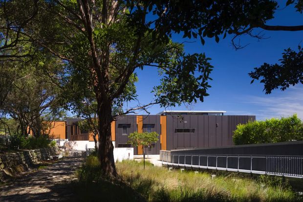 Exterior view of a contemporary, low-profile building set within a leafy landscaped site. The facade combines dark vertical panel cladding with warm orange-brown feature sections and broad horizontal glazing. A paved pedestrian path, mature trees, grasses, retaining walls, and a dark metal-railed bridge or access structure occupy the foreground.
