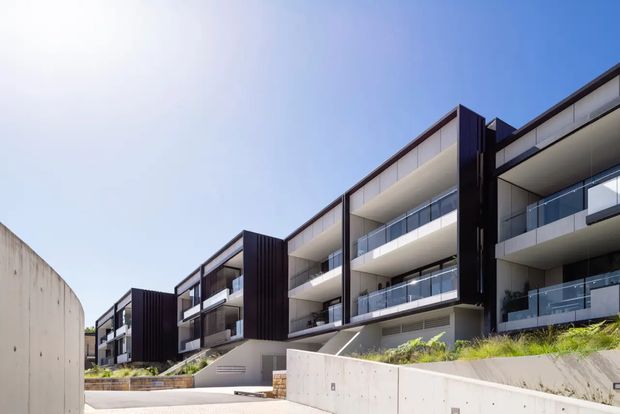 Contemporary multi-storey apartment building viewed from outside, featuring stepped massing, deep balconies with clear glass balustrades, dark vertical facade panels, pale rendered or concrete walls, and landscaped terraces. Bright blue sky and strong sunlight emphasize the clean modern geometry.
