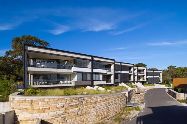 Contemporary multi-storey residential development viewed from a curved driveway, featuring long horizontal wings, dark-framed floor-to-ceiling glazing, projecting balconies, white and grey facade panels, and landscaped terraced stone retaining walls. The complex sits in a wooded suburban setting beneath a clear blue sky.