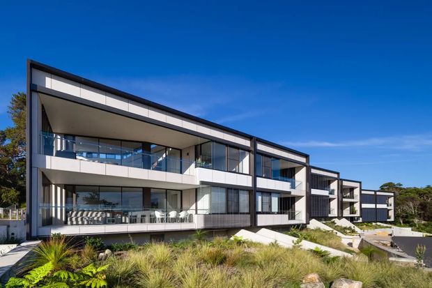 Exterior view of a contemporary multi-storey residential building with strong horizontal lines, extensive floor-to-ceiling glazing, projecting balconies, dark structural framing, and light panel cladding. The building is set within landscaped coastal-style planting under a clear blue sky.