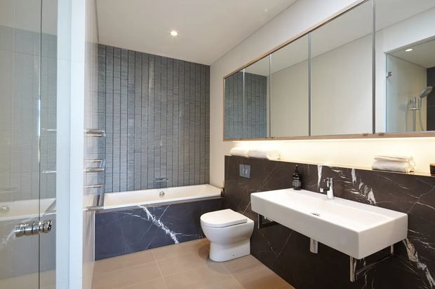 Contemporary bathroom with a freestanding-style bathtub set against a dark marble-look tiled feature wall, a wall-mounted white vanity basin, toilet, large mirrored cabinetry, and a glass shower enclosure. Neutral beige floor tiles, recessed ceiling lighting, and timber-toned trim create a refined, high-contrast interior.