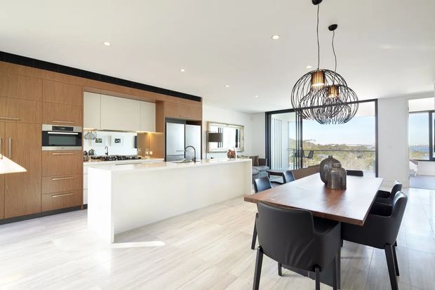 Open-plan contemporary kitchen and dining area with a large white island, integrated timber cabinetry, stainless-steel appliances, and a timber dining table with dark upholstered chairs. Full-height glazing opens toward a bright outdoor view, while two oversized black wire pendant lights define the dining zone.