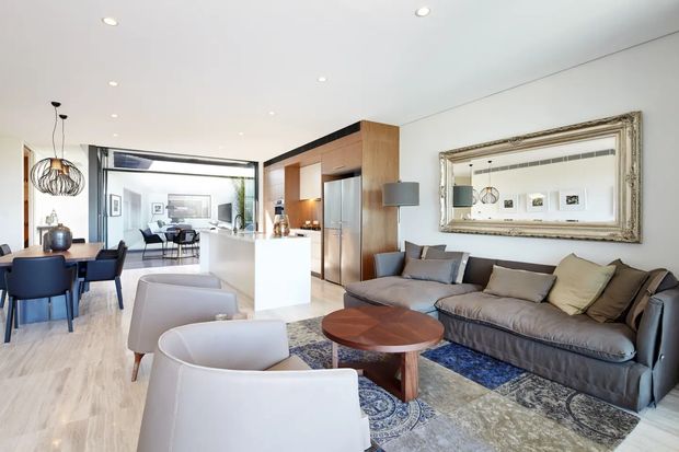 Bright open-plan contemporary living and dining area with a compact kitchen. The room features pale timber flooring, a white island, warm timber cabinetry, a grey sectional sofa, sculptural pendant lighting, blue dining chairs, a patterned area rug, and a large ornate wall mirror. Glazing at the rear opens the space toward another seating area and greenery.