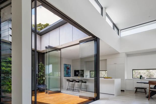 Bright contemporary open-plan interior viewed through a large glazed opening, with a compact white kitchen, island bench and stools, dining area, polished light flooring, and a timber-floored courtyard or sunroom separated by frameless glass sliding doors. The space features strong natural light, white surfaces, dark slim window frames, and a high ceiling with clerestory glazing.