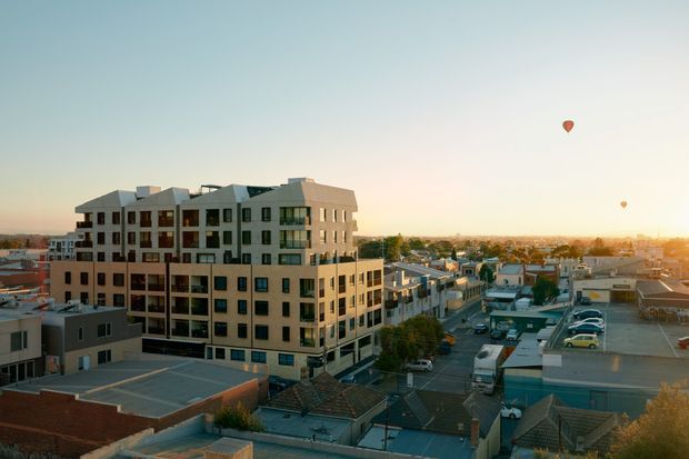 Aerial view of a contemporary multi-storey apartment building in an urban suburban setting. The building has a stepped massing, pale masonry or concrete facade, extensive dark-framed windows and balconies, and a varied pitched roofline. Low-rise commercial and residential buildings, streets and parked vehicles surround it, with hot-air balloons visible in the distant sky.